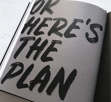 Bold black brush lettering on a planner page reading “OK HERE’S THE PLAN” — a powerful cue to get organised and take action.