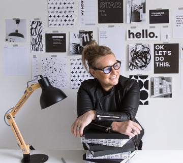 Woman smiling at her desk surrounded by HELLO TIME planners and a creative wall of monochrome inspiration prints and motivational quotes.