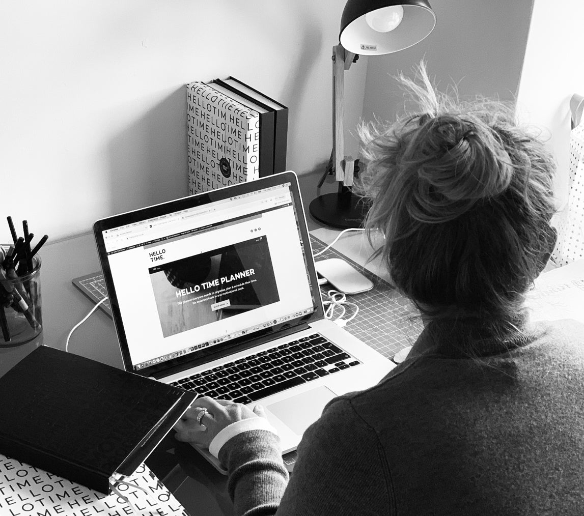 Woman working at her desk with the HELLO TIME website open on a laptop, surrounded by planners, notebooks, and creative workspace energy.