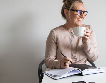 Woman smiling while holding a mug and planning her day with a HELLO TIME planner at a white desk. Calm, focused, and ready to go.