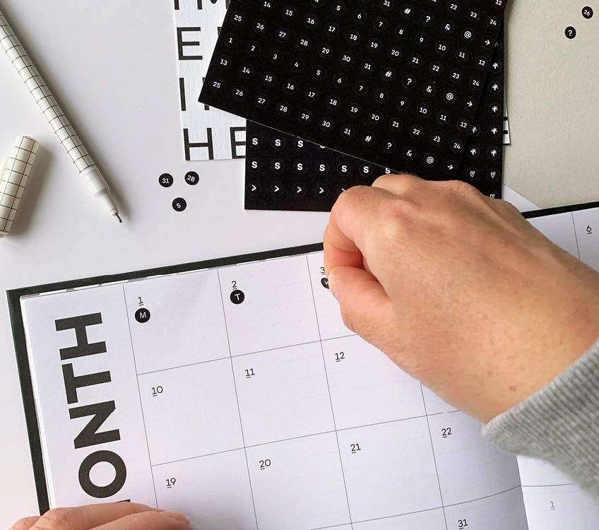 Hand placing black date stickers onto a minimalist monthly planner page, surrounded by pens and stationery for organised planning.