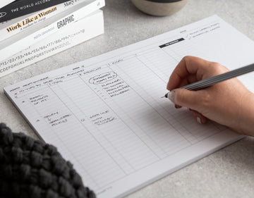 Hand writing in a HELLO TIME weekly planner pad on a grey desk, surrounded by books — planning goals, priorities and creative ideas.
