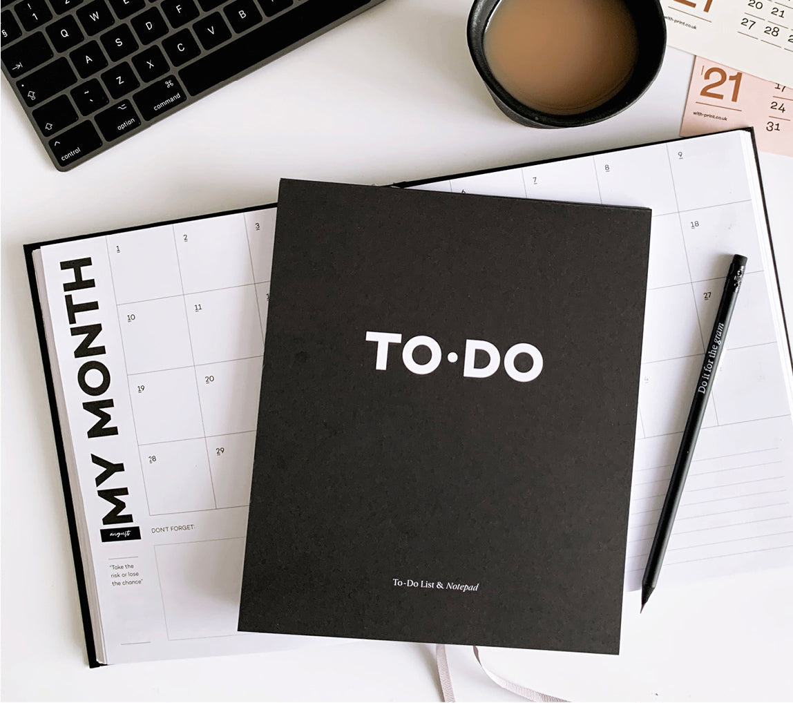Black TO·DO notepad on top of an open monthly planner, with pencil, coffee cup, and keyboard — minimalist desk setup for getting things done.