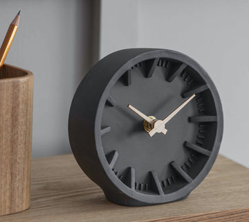 Minimalist dark grey desk clock with wooden hands, sitting on a light wood surface beside a pencil pot — calm, modern, and perfectly on time.