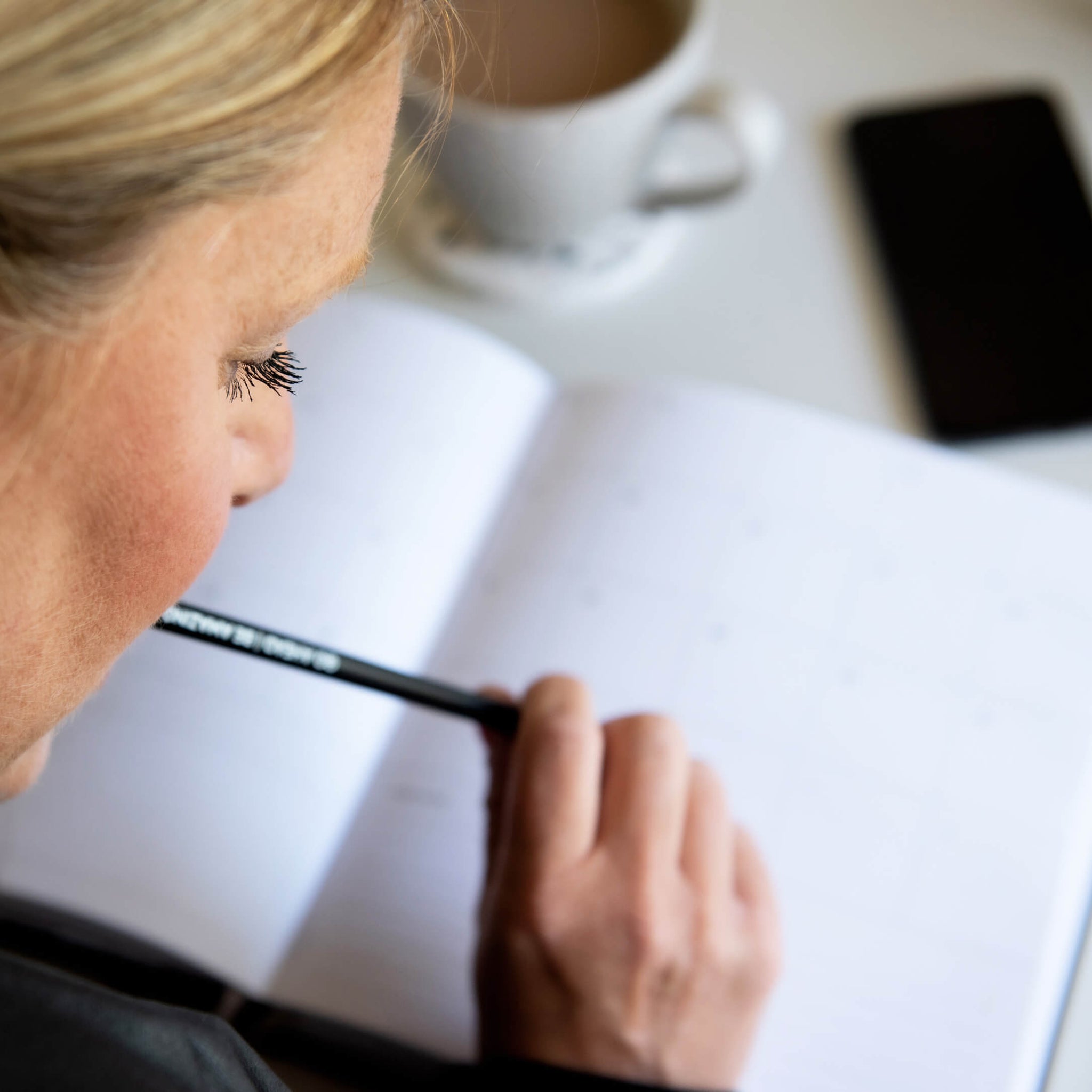 Woman planning in an open HELLO TIME planner with pencil in hand, phone and coffee nearby — focused on organising her week with intent.