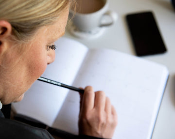 Woman planning in an open HELLO TIME planner with pencil in hand, phone and coffee nearby — focused on organising her week with intent.