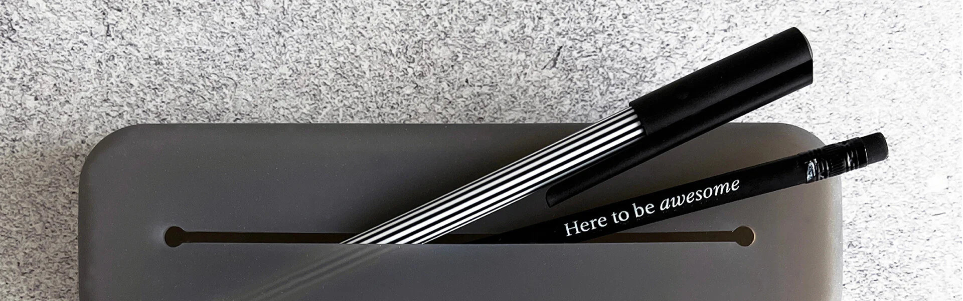 Two black pens, one with "Here to be awesome" text, resting on grey case against textured grey surface