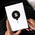 White children’s birthday card with black balloon and letter S initial, held in hands.