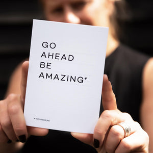 Person holding white “Go Ahead Be Amazing” card with bold black typography.