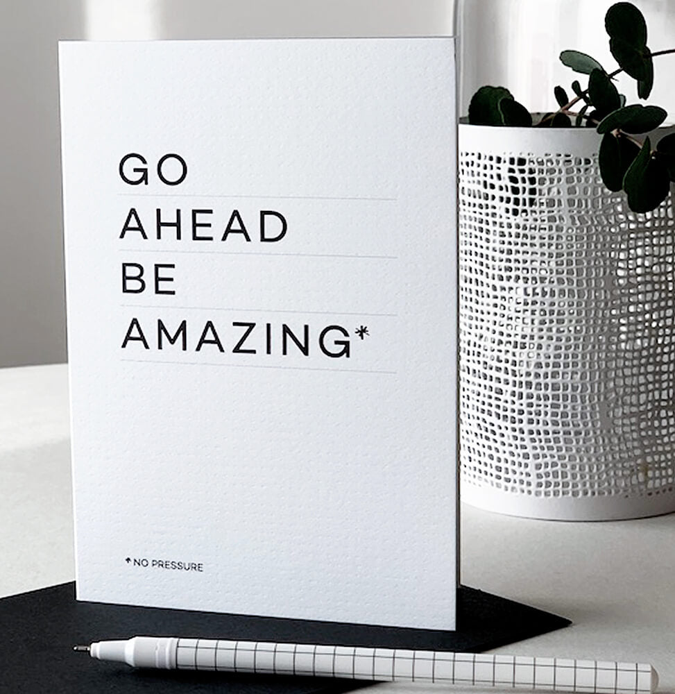 White “Go Ahead Be Amazing” card displayed upright beside pen and decorative plant.
