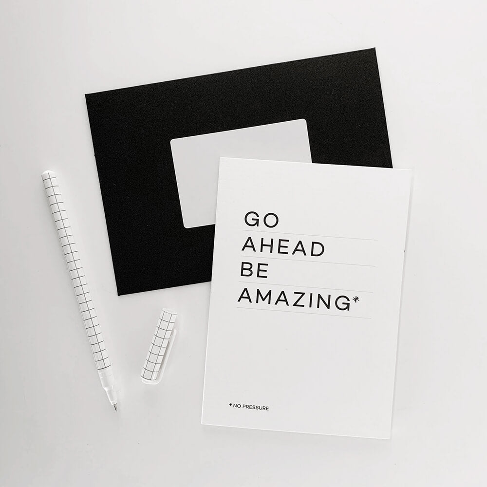 Monochrome greetings card reading “Go Ahead Be Amazing” with black envelope and pen.