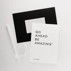 Monochrome greetings card reading “Go Ahead Be Amazing” with black envelope and pen.