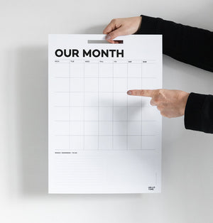 Hands holding and pointing at undated monthly Hello Time wall planner on white wall