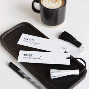 “ON THE SAME PAGE” and “TO BE CONTINUED” bookmarks with leather tassels next to coffee and pen