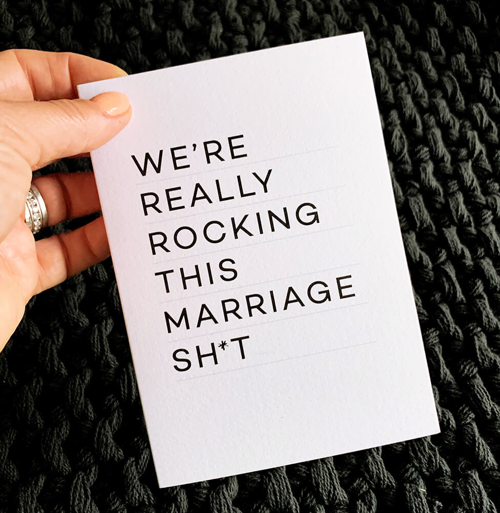 Hand holding monochrome anniversary card with bold text "We’re really rocking this marriage sh*t."