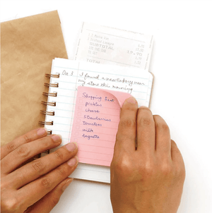 Hand placing a pink to-do sticky tab on a small notebook, shopping list written, receipt tucked behind page.