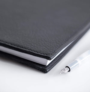 Close-up of HELLO TIME black leather notebook cover with white pages and a transparent Kaweco fountain pen.