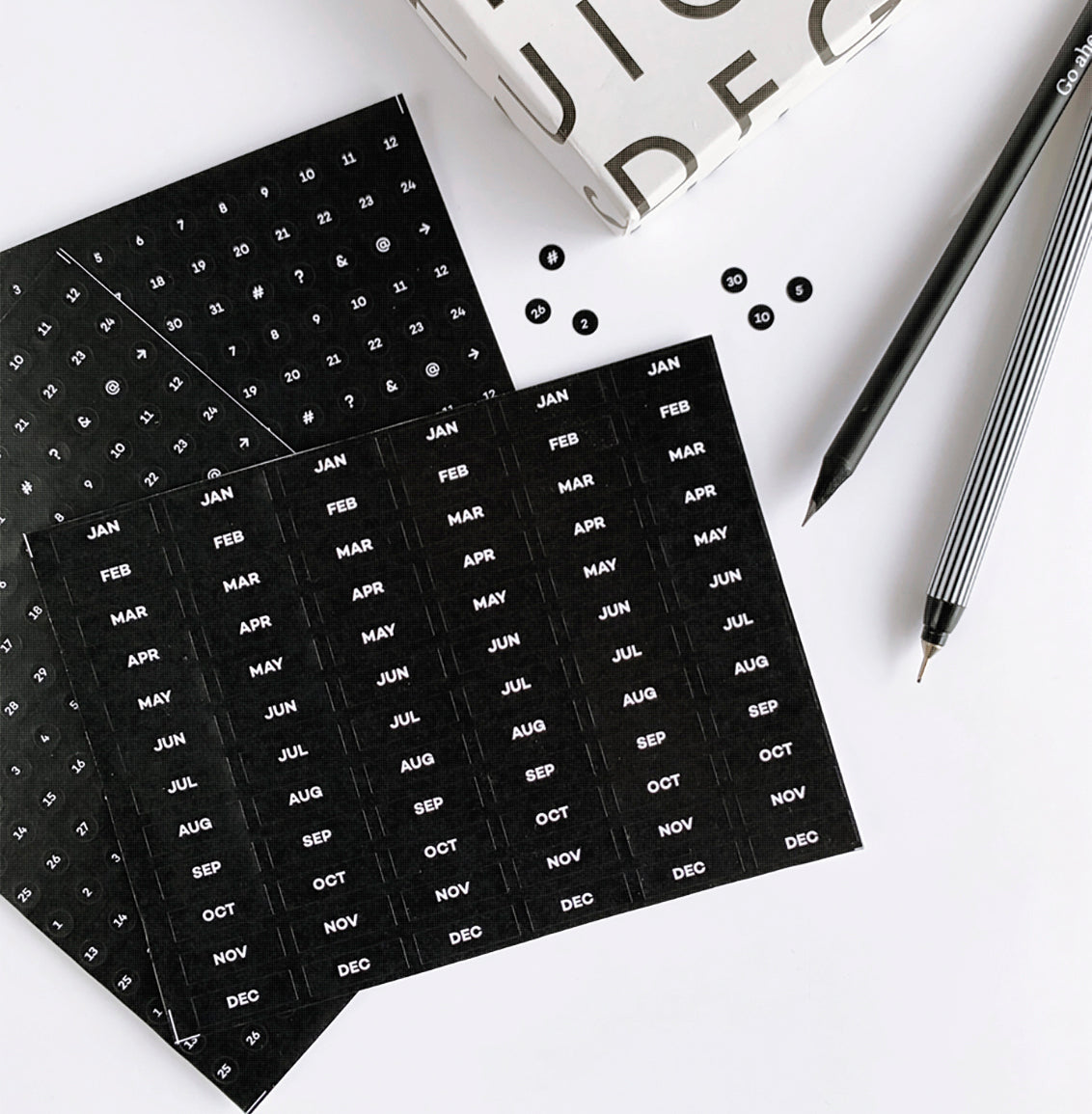Two black HELLO TIME diary sticker sheets with white months and dates, shown with pens and monochrome packaging