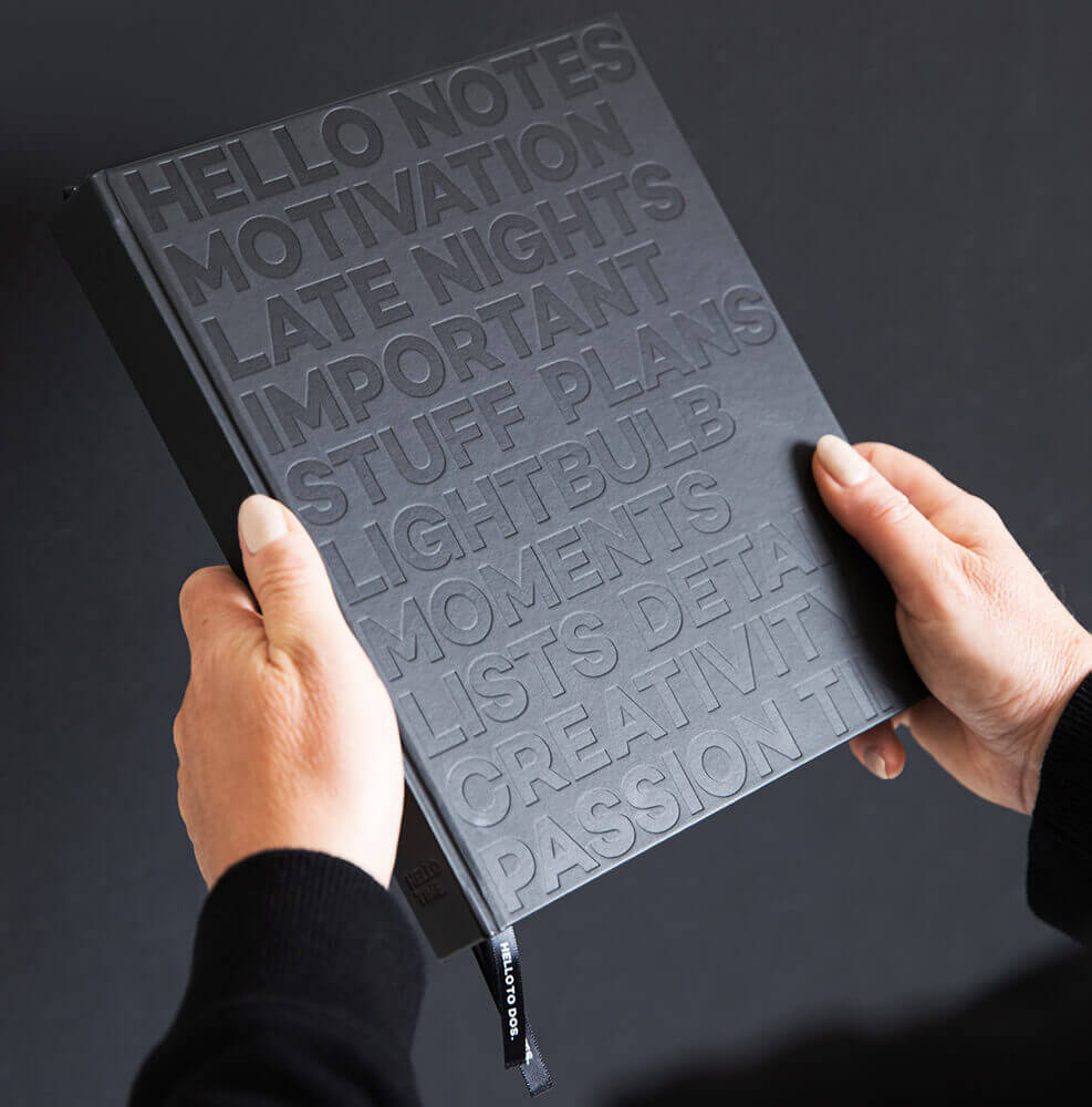 Hands holding black HELLO TIME notebook with embossed motivational words on matte cover