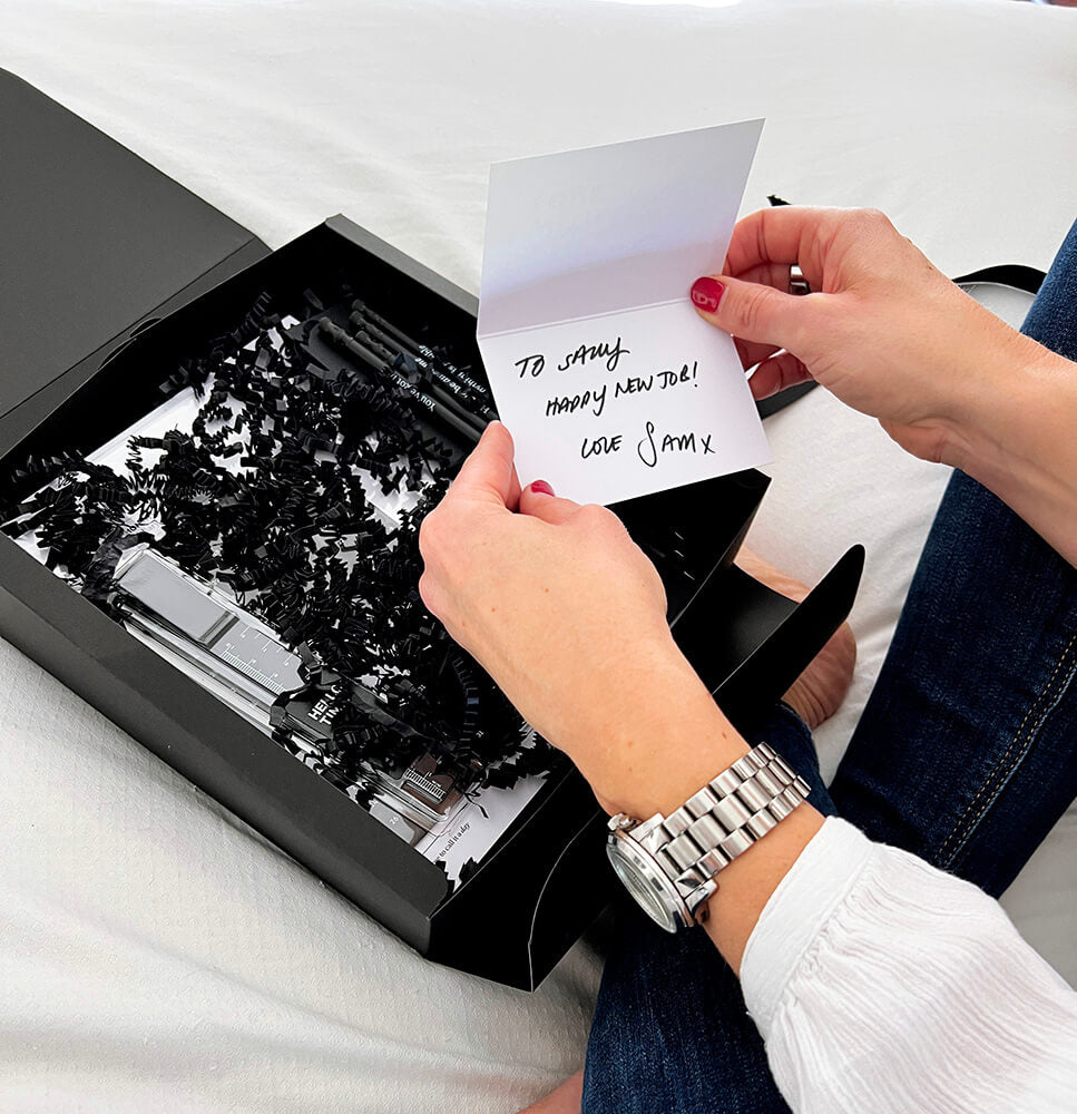 Person opening black HELLO TIME stationery gift box with handwritten new gift card and black shredded filler