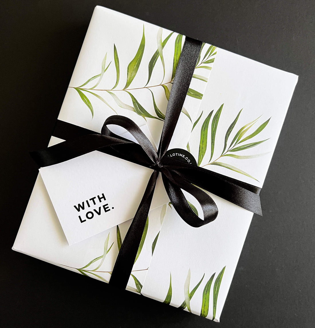 HELLO TIME gift-wrapped box in fern print paper with black ribbon and white tag reading “WITH LOVE.” on black background