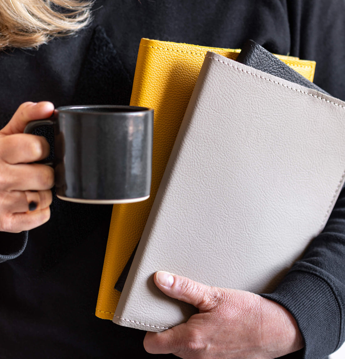 Close-up of a woman holding three HELLO TIME Luxe Leather Notebook Covers in yellow, grey, and black, along with a black coffee mug