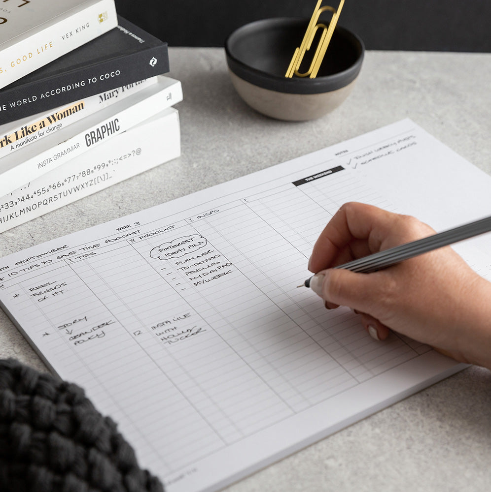 Hand writing on HELLO TIME My Week desk pad with books and paperclips on desk