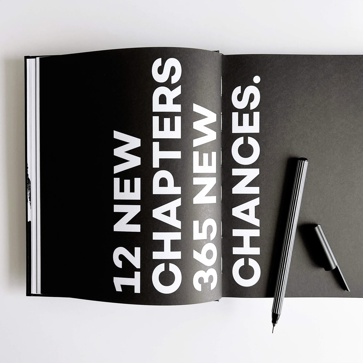 Open black HELLO TIME planner spread with white "12 new chapters, 365 new chances" text with a black and white striped pen