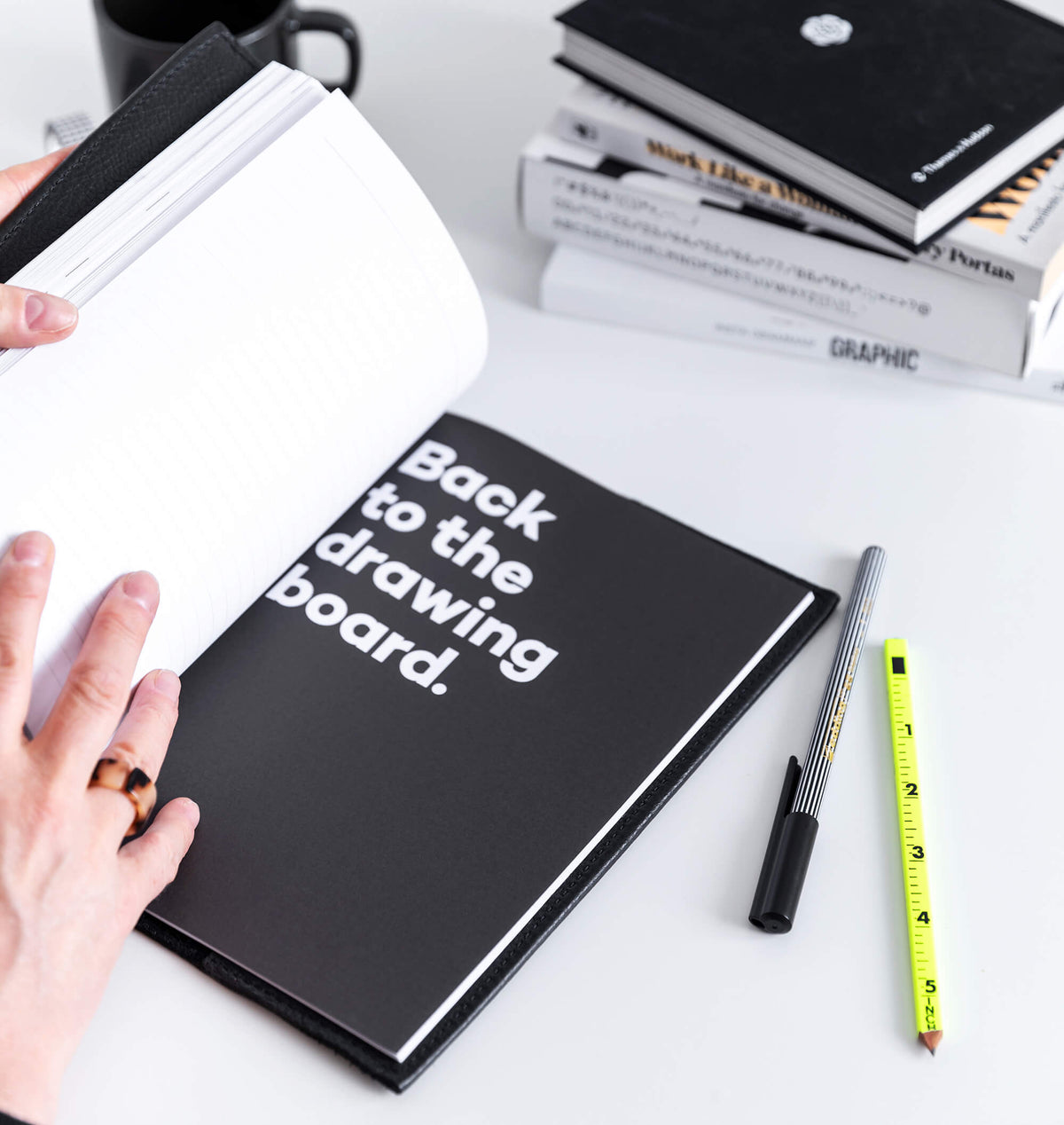 Open black HELLO TIME notebook with "Back to the drawing board" text, hand flipping pages, pencil and pen beside it.