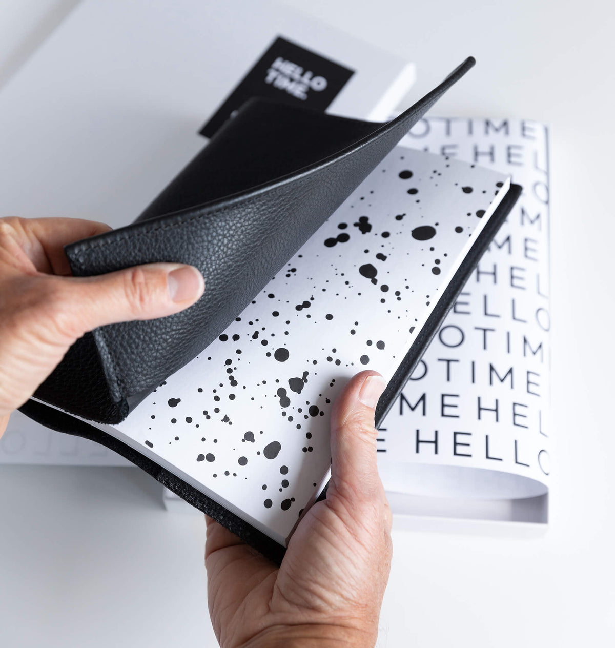 Hands opening black leather HELLO TIME planner cover over inkwell endpaper, HELLO TIME branded box behind.