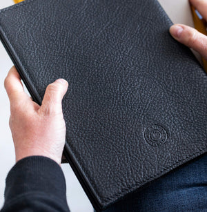Hands holding closed black leather HELLO TIME planner cover with embossed logo detail.