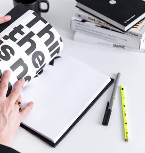 Hand opening black HELLO TIME notebook to blank page, bold black typography tab visible, pen and pencil nearby.