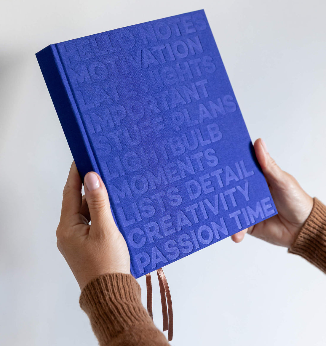 Hands holding bright blue HELLO TIME planner with embossed cover text and ribbon bookmarks hanging below.