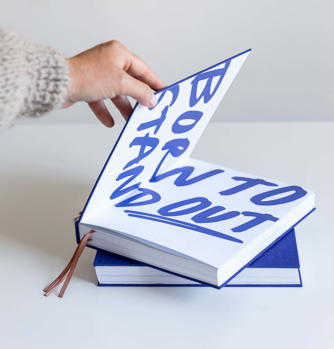 Hands holding bright blue HELLO TIME planner with embossed cover text and ribbon bookmarks hanging below.