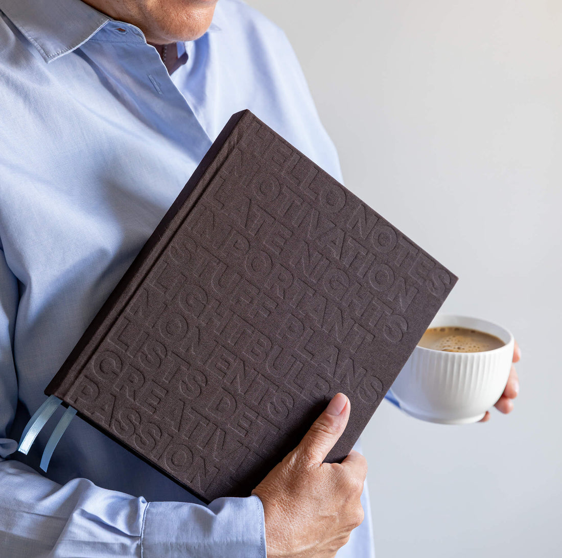 Person in blue shirt holding brown HELLO TIME planner with embossed cover and pale blue ribbons, coffee cup in other hand.
