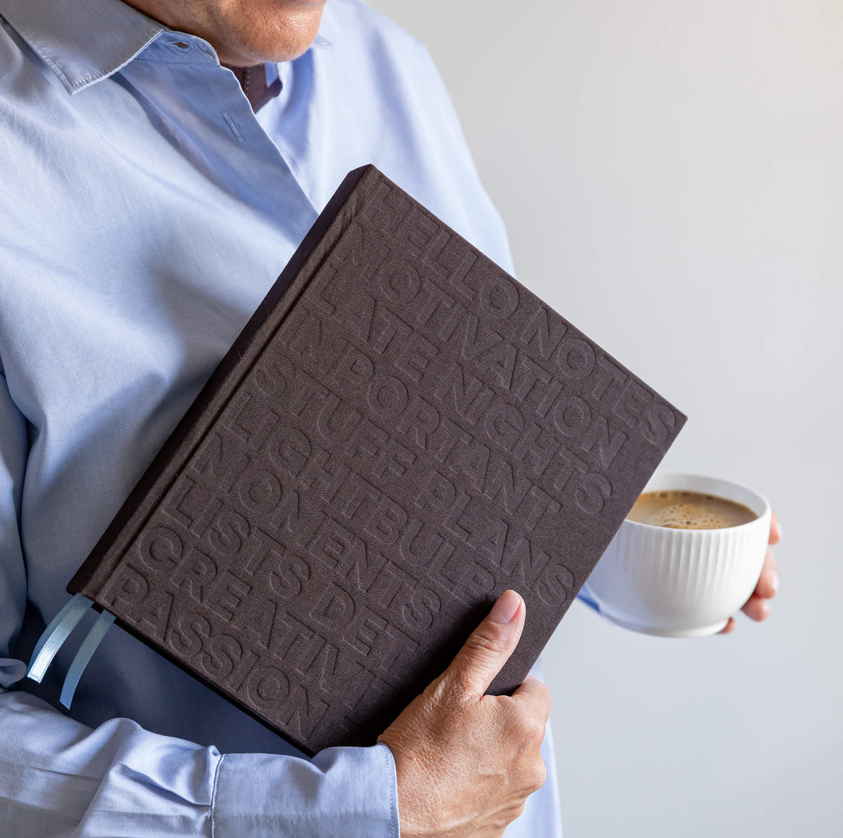Person in blue shirt holding brown HELLO TIME planner with embossed cover and pale blue ribbons, coffee cup in other hand.