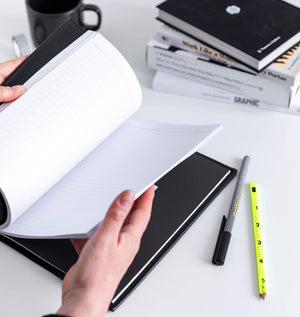 Hands flipping through black HELLO TIME notebook's lined pages, pen, pencil and books on desk behind.