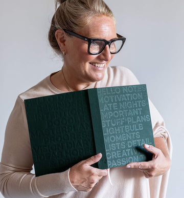 Smiling woman in glasses holding open green HELLO TIME planner showing embossed text on front and back covers.