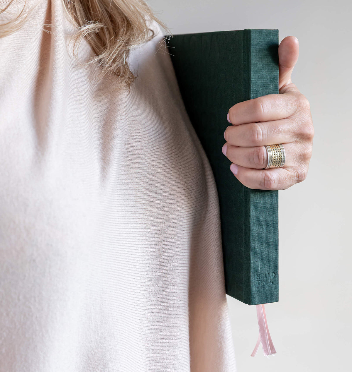 Person in cream jumper holding green HELLO TIME planner against their side, ribbon bookmarks hanging below.