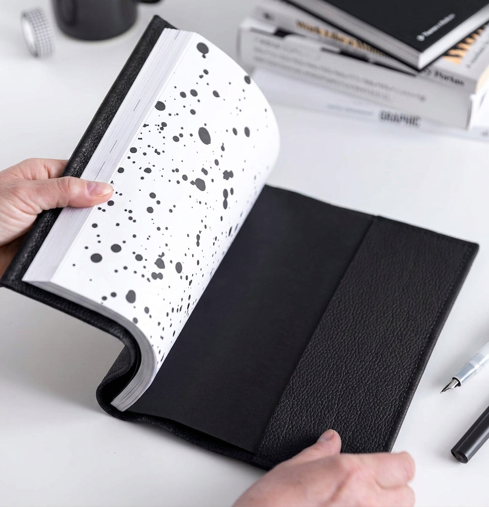 Hands opening black leather HELLO TIME notebook cover with black inkwell endpaper, pen beside it.