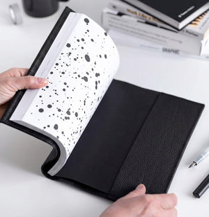 Hands opening black leather HELLO TIME notebook cover with black inkwell endpaper, pen beside it.