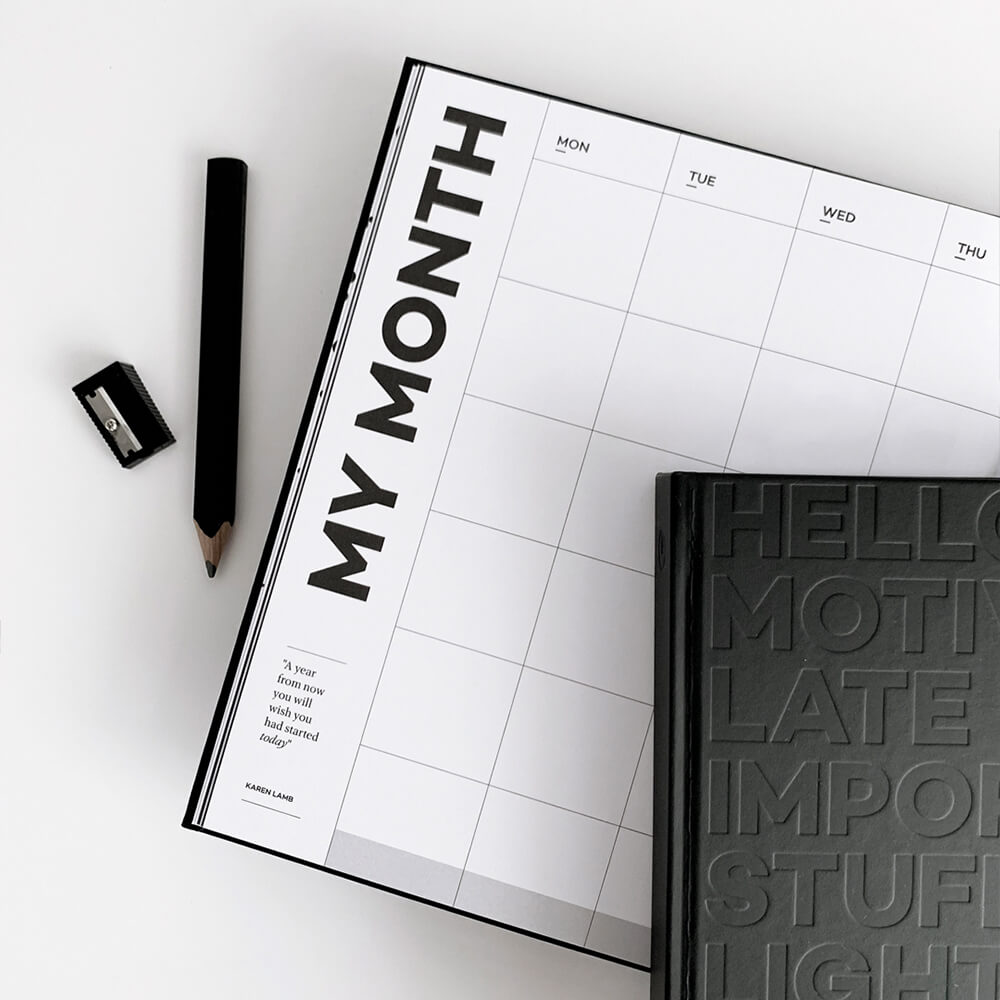 Open HELLO TIME "My Month" planner page with weekly grid, black embossed planner cover, pencil and sharpener beside it.