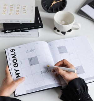 HELLO TIME planner open to My Goals page with hand writing in grid layout, surrounded by books, pen, coffee cup and black desk clock