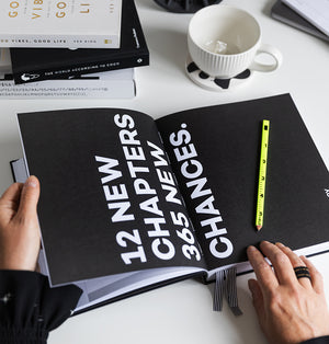 HELLO TIME planner open to bold black page with white text reading “12 new chapters, 365 new chances” with hands and neon pencil on desk