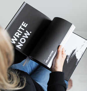 Person holding HELLO TIME planner open to black “WRITE NOW” and “HELLO NOTES” section with bold typography and monochrome design