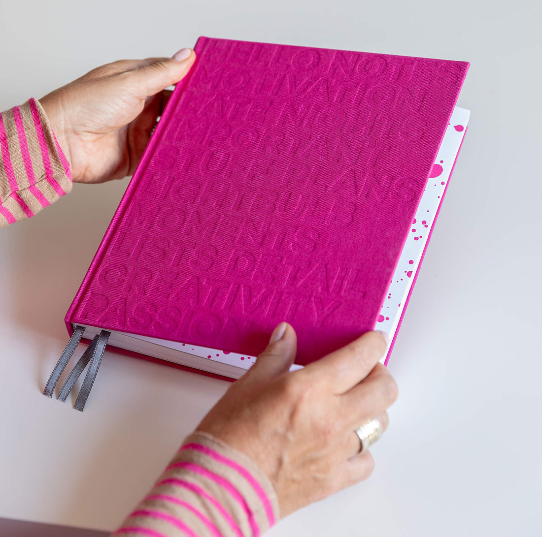 Hands holding closed pink HELLO TIME planner with embossed cover text, ribbon bookmarks and pink inkwell endpaper.