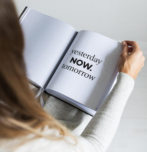Person holding HELLO TIME planner open to white page with bold text reading “yesterday, NOW, tomorrow” and striped ribbon markers visible