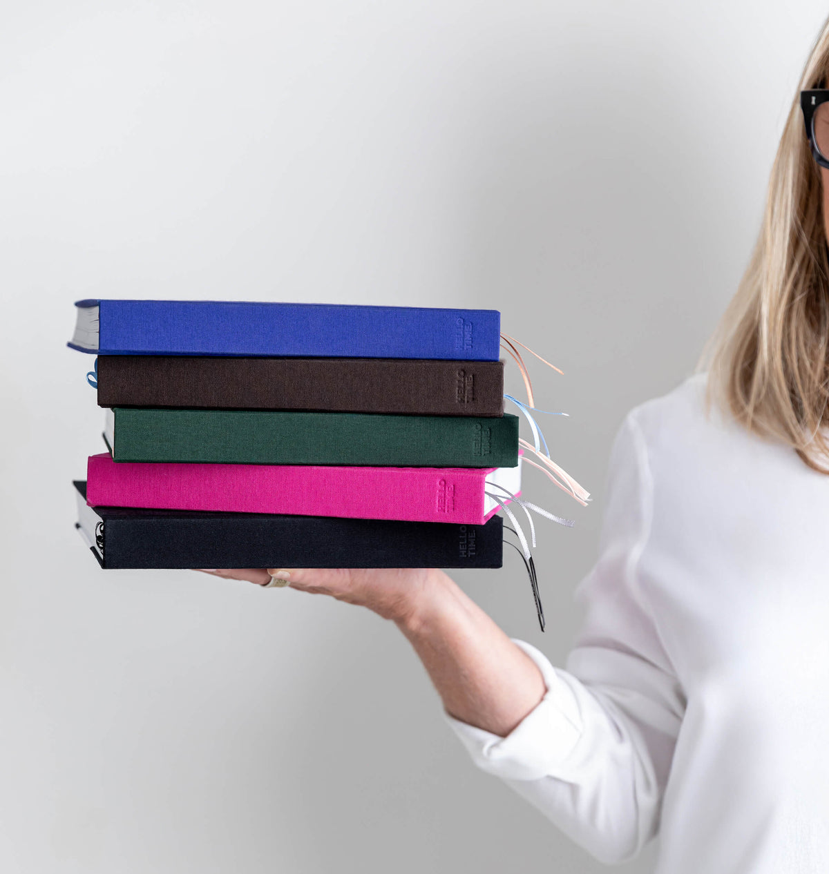 Woman holding a stack of five HELLO TIME planners in blue, brown, green, pink and black with ribbon bookmarks showing.