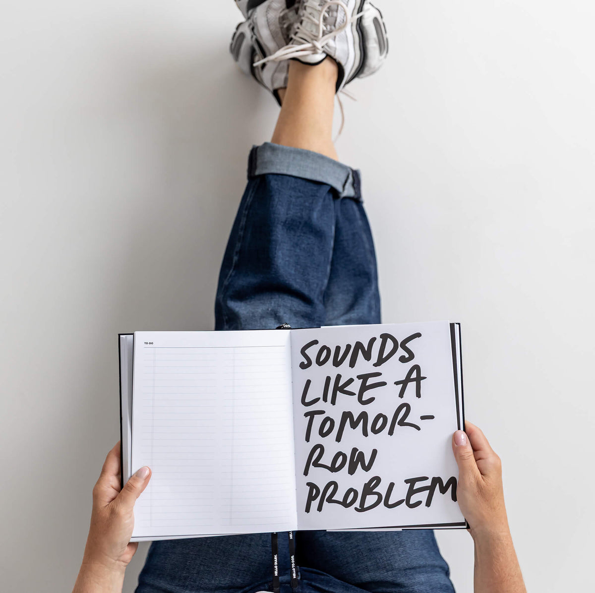 Person lying back with feet up a wall, holding open HELLO TIME planner with handwritten “Sounds like a tomorrow problem” page.