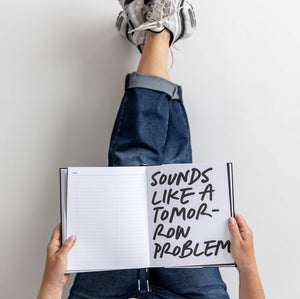 Person lying back with feet up a wall, holding open HELLO TIME planner with handwritten “Sounds like a tomorrow problem” page.